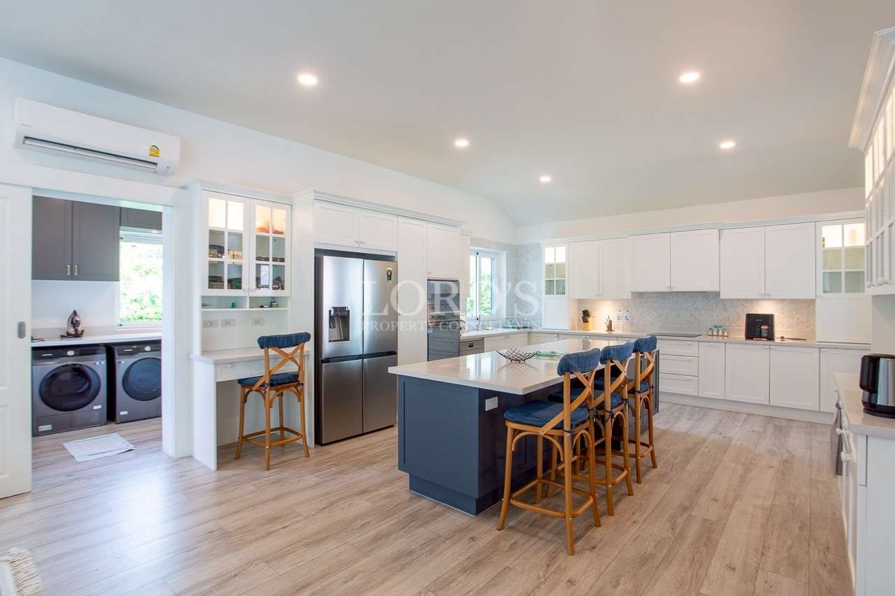 Open-plan kitchen with blue island, white cabinets, bar stools, stainless steel fridge, and adjacent laundry room.