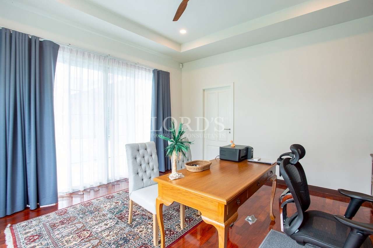 Home office with wooden desk, office chair, printer, large window with curtains, and patterned rug.