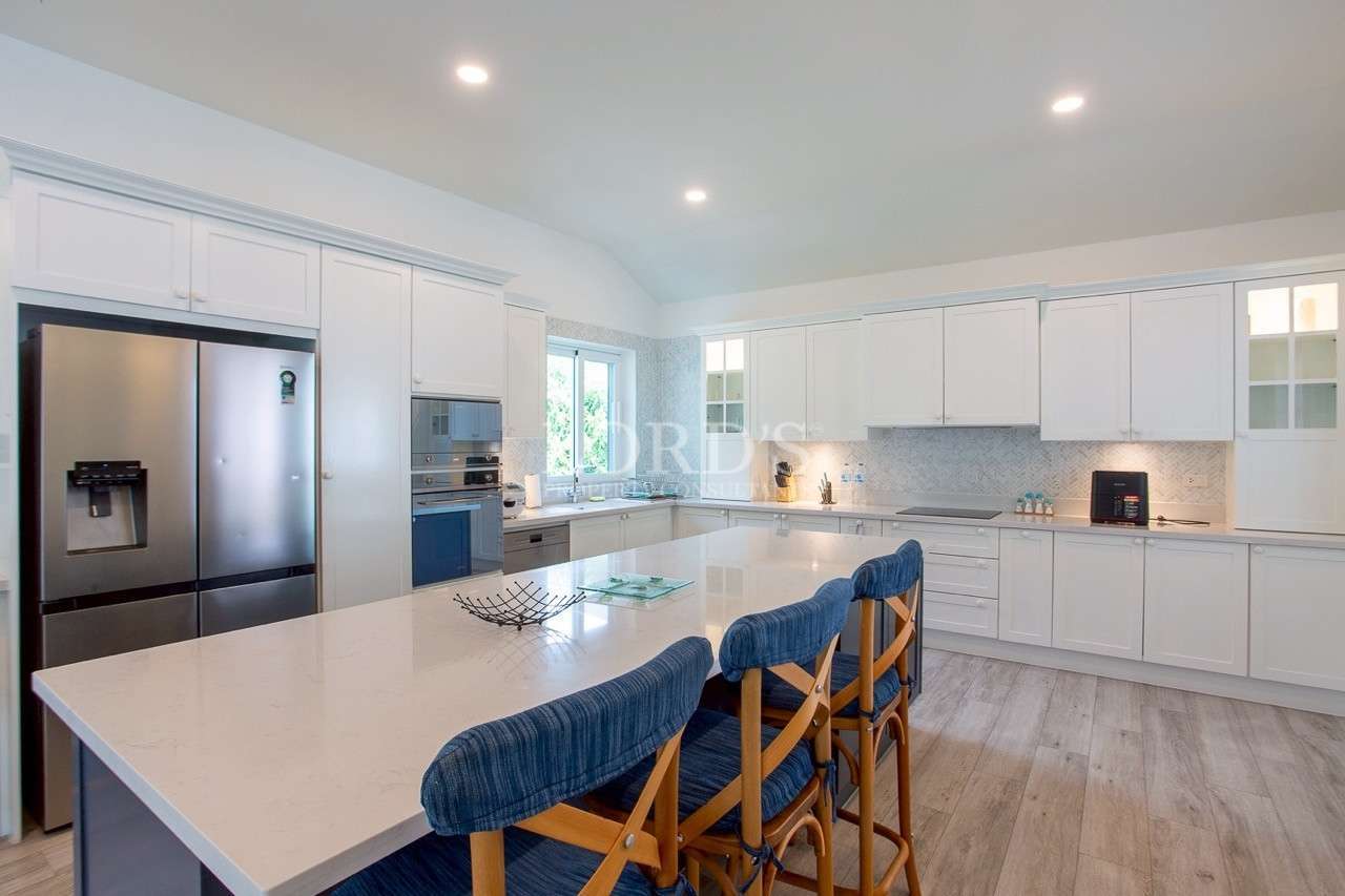 Modern white kitchen with large island, blue bar stools, stainless steel refrigerator, and built-in appliances.