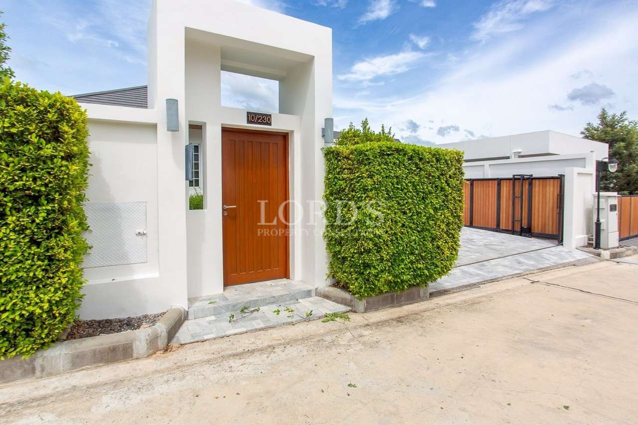 Modern villa entrance with wooden gate, white facade, and trimmed hedge wall.