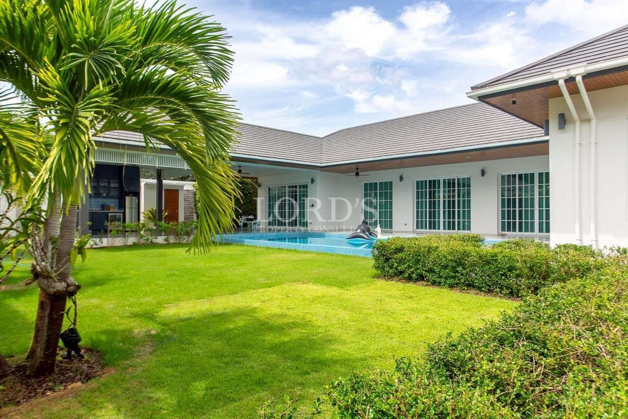 Modern white villa with large sliding glass doors, private swimming pool, and lush green lawn with a palm tree.