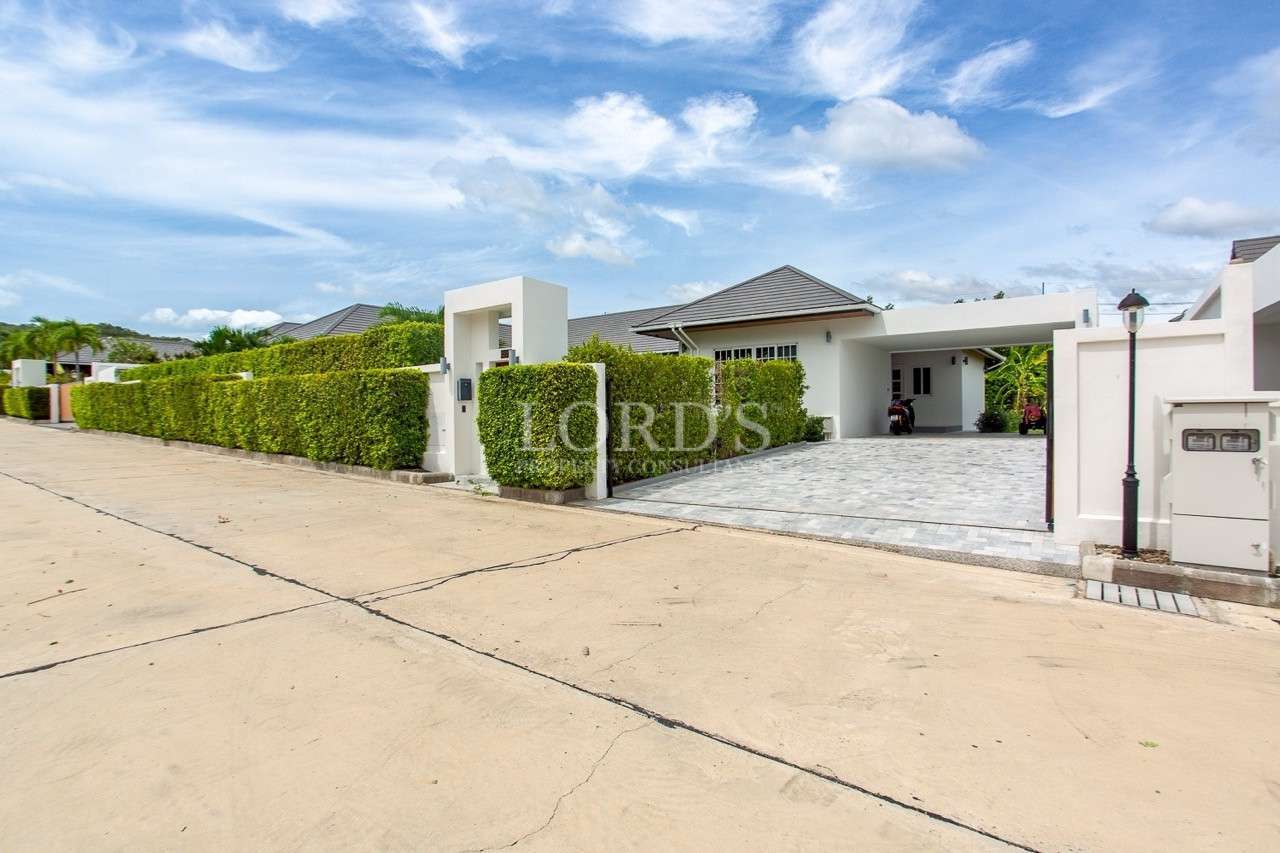 Modern single-story white villa with gated entrance, driveway, and tall green hedges under a bright blue sky.