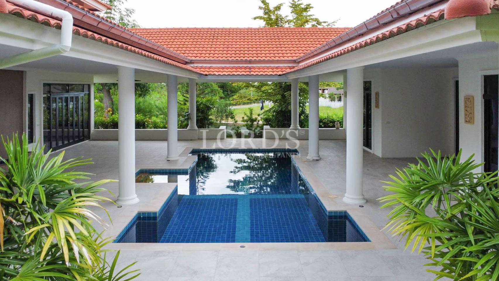 Covered courtyard pool with blue tiles, white columns, and red-tiled roof in a tropical villa.