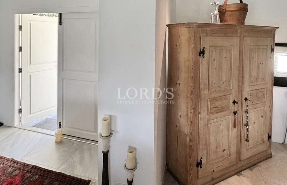 Rustic wooden cabinet with decorative hardware beside a white doorway and candle stands in a minimalist interior.