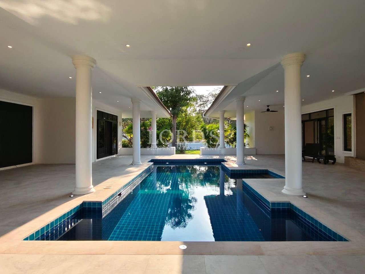 Luxury villa courtyard with covered patio, white columns, and central blue tiled swimming pool surrounded by tropical greenery.