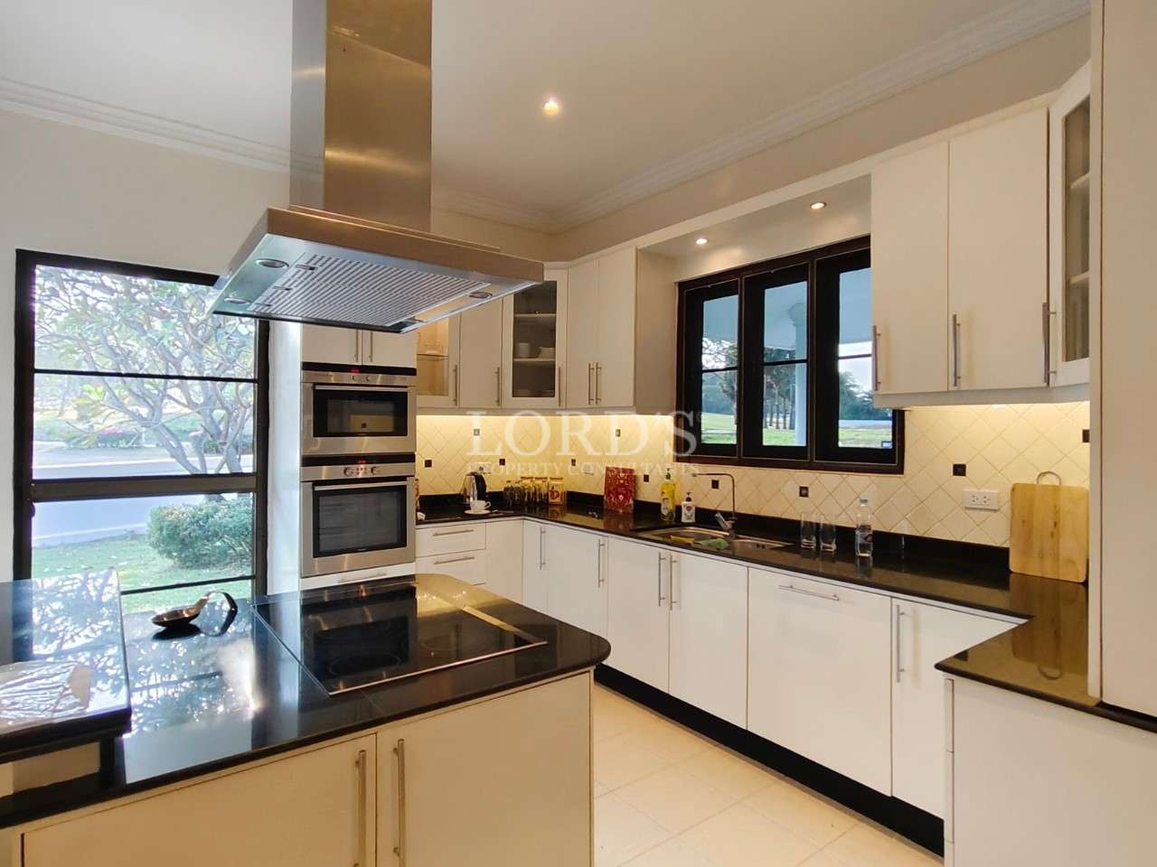Modern white kitchen with black granite countertops, built-in oven, island cooktop, and large windows with garden view.