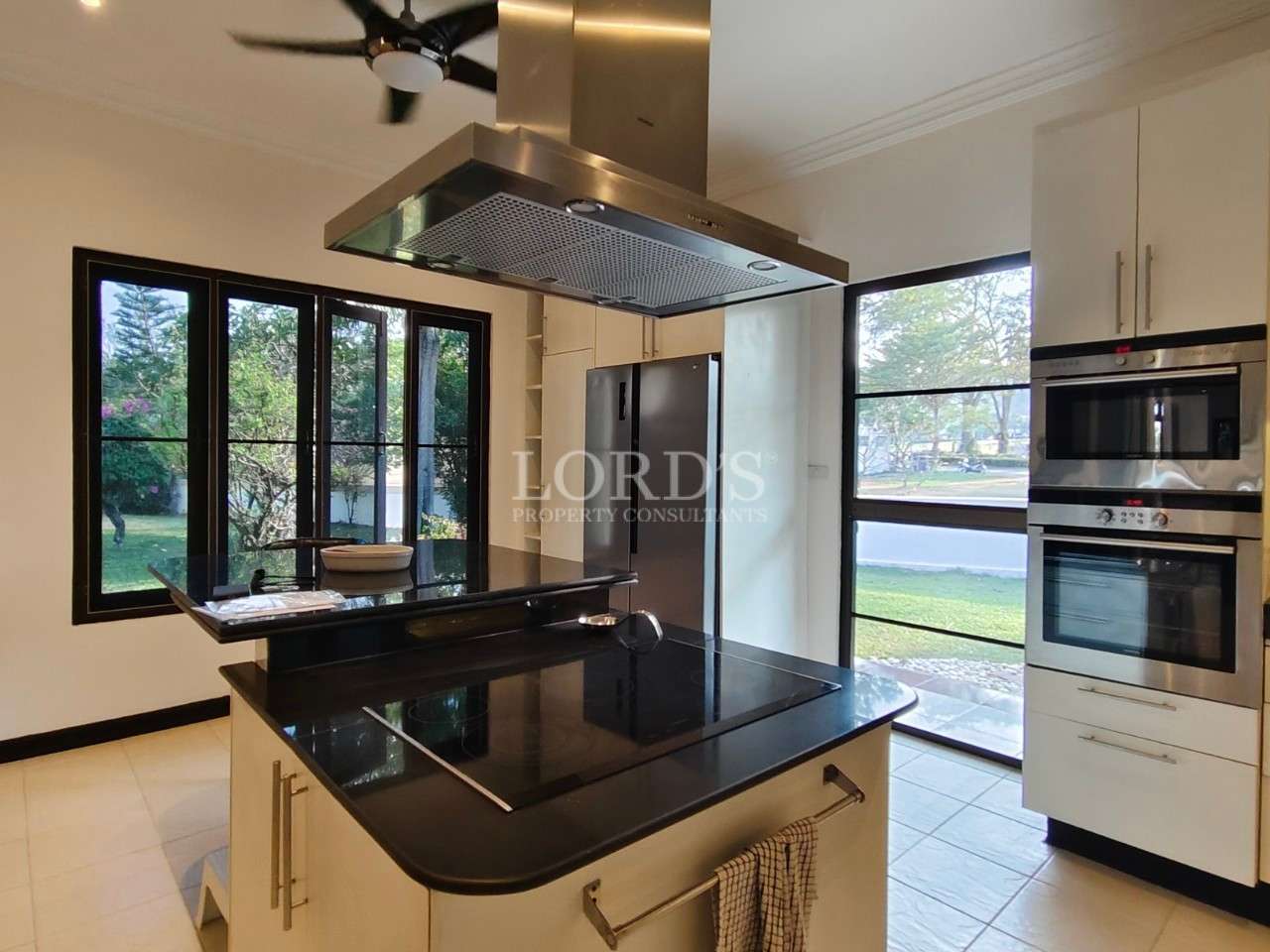 Modern kitchen with black island cooktop, stainless steel range hood, built-in oven, and large windows overlooking the garden.