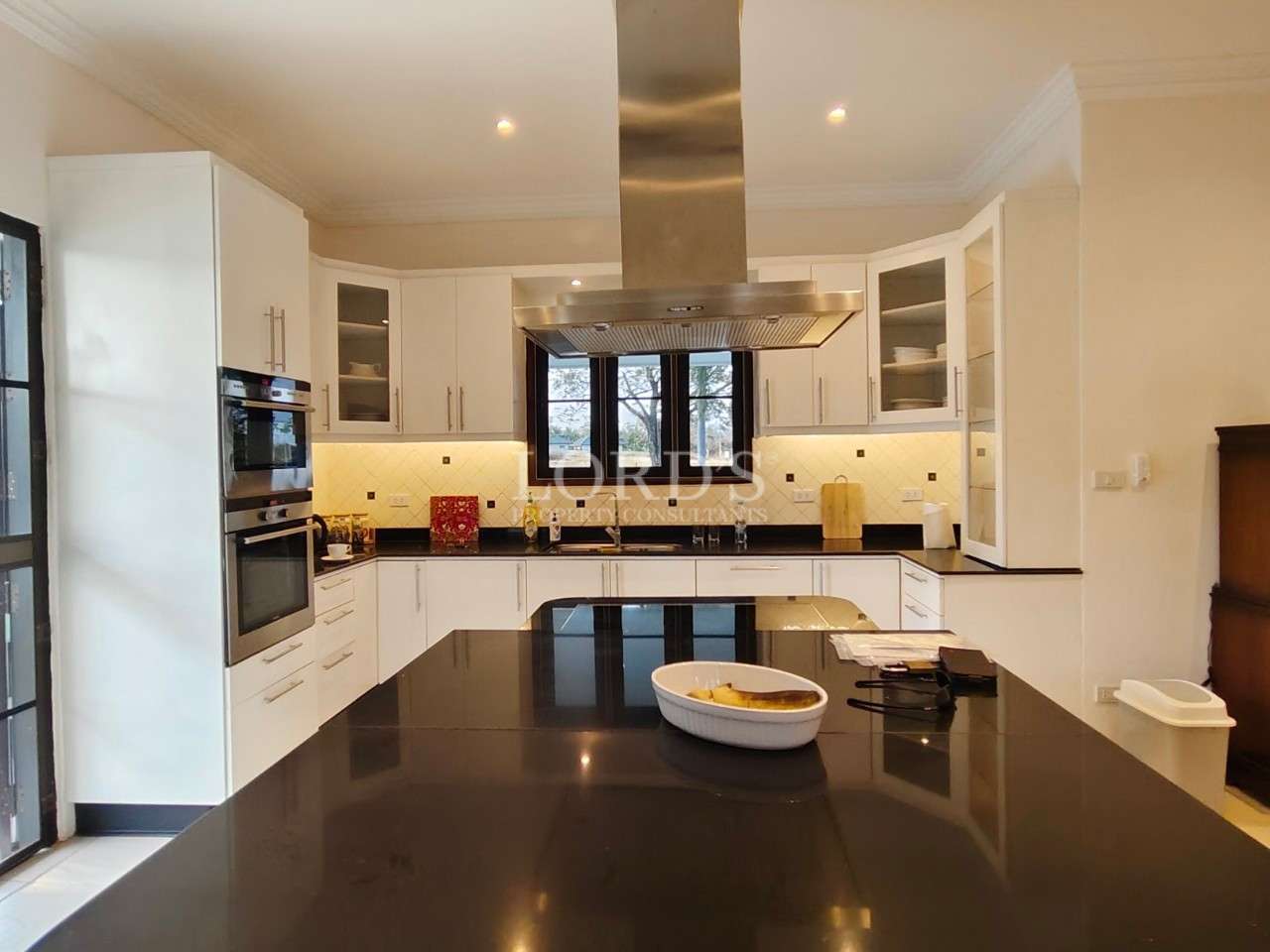 Modern white kitchen with black island countertop, built-in oven, stainless steel range hood, and under-cabinet lighting.