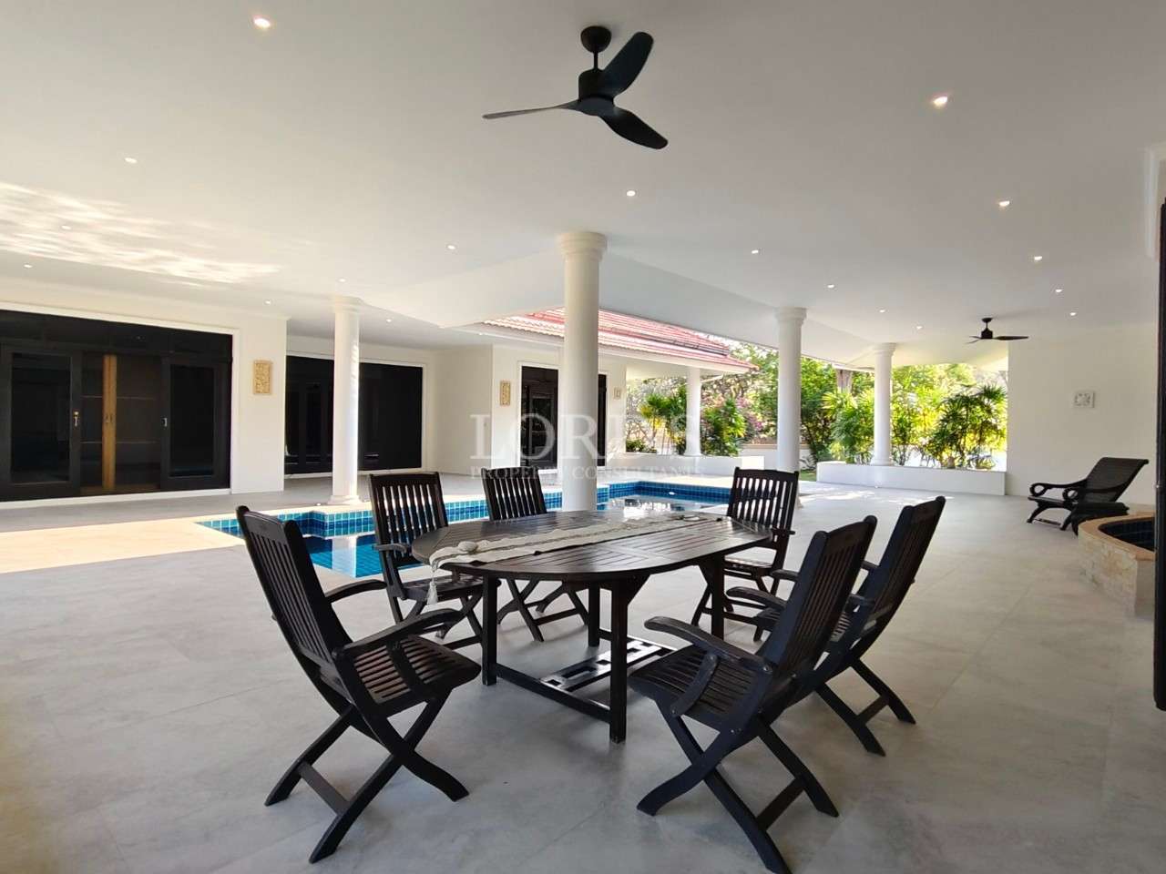 Spacious covered terrace with wooden outdoor dining set, ceiling fans, white columns, and pool view.