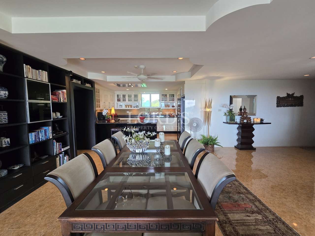 Spacious dining room with glass-top table, upholstered chairs, built-in shelving, and open kitchen in the background.