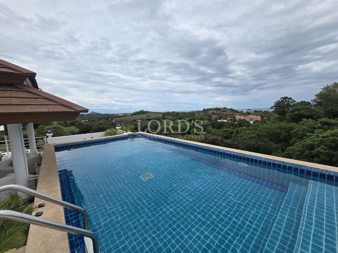 Infinity swimming pool with panoramic views of hills, greenery, and distant coastline under a cloudy sky.