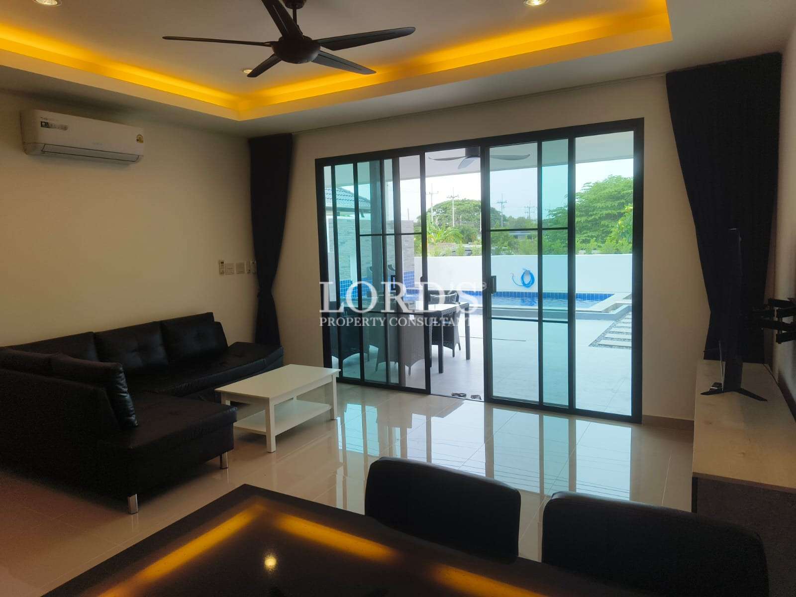 Modern living room with black sofa, white coffee table, ceiling fan, and large sliding glass doors opening to a private pool area.