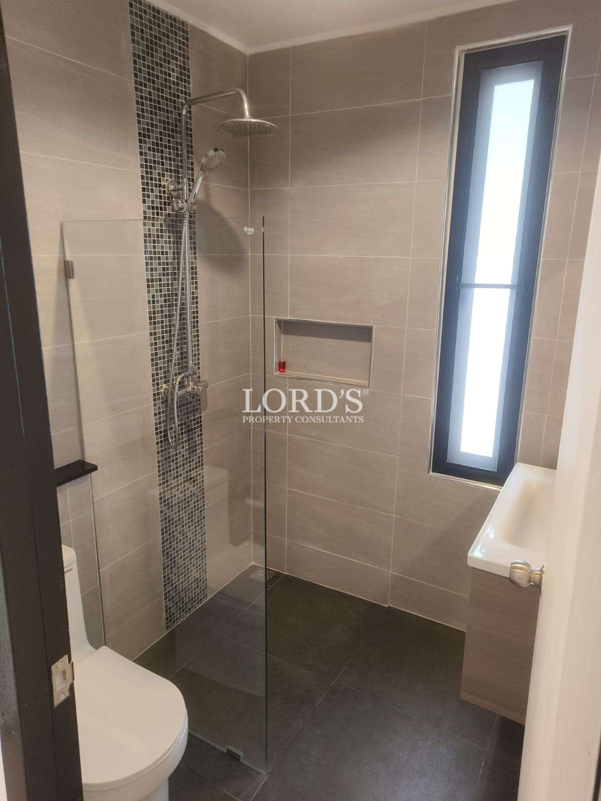 A compact contemporary bathroom featuring a glass-enclosed walk-in shower with a rain showerhead and handheld fixture. A vertical strip of mosaic tiles adds a decorative accent behind the shower. The space includes gray wall tiles, dark floor tiles, a built-in wall niche for toiletries, a white toilet, and a small wall-mounted sink beside a tall frosted window that allows natural light while maintaining privacy.