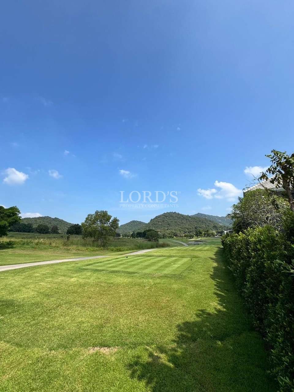 Manicured golf fairway with striped grass, bordered by hedges and trees, with rolling hills under a bright blue sky.
