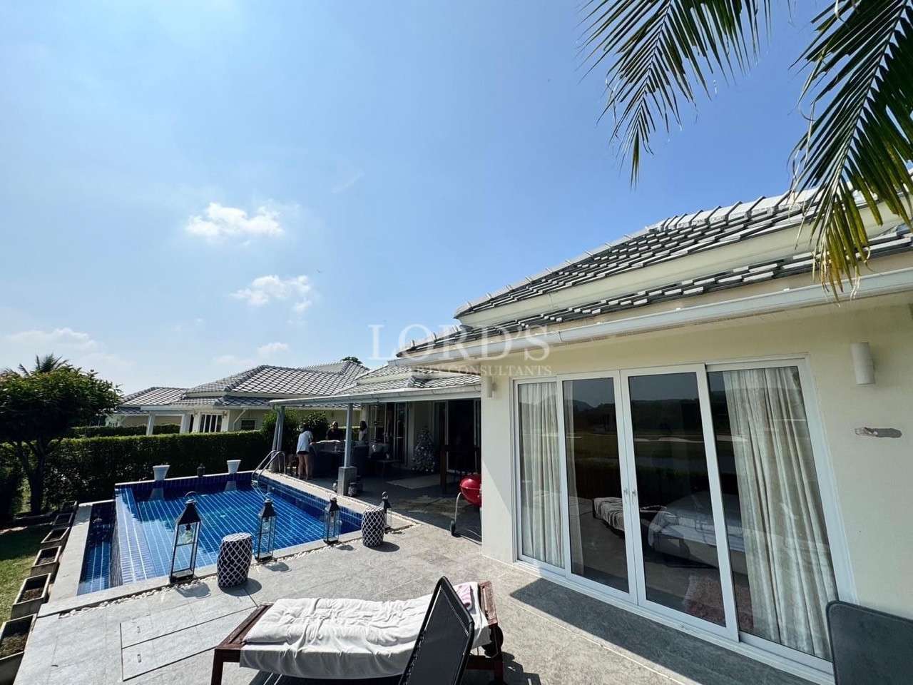 Modern single-story villa with sliding glass doors, tiled roof, and a private blue swimming pool with lounge chairs under a sunny sky.