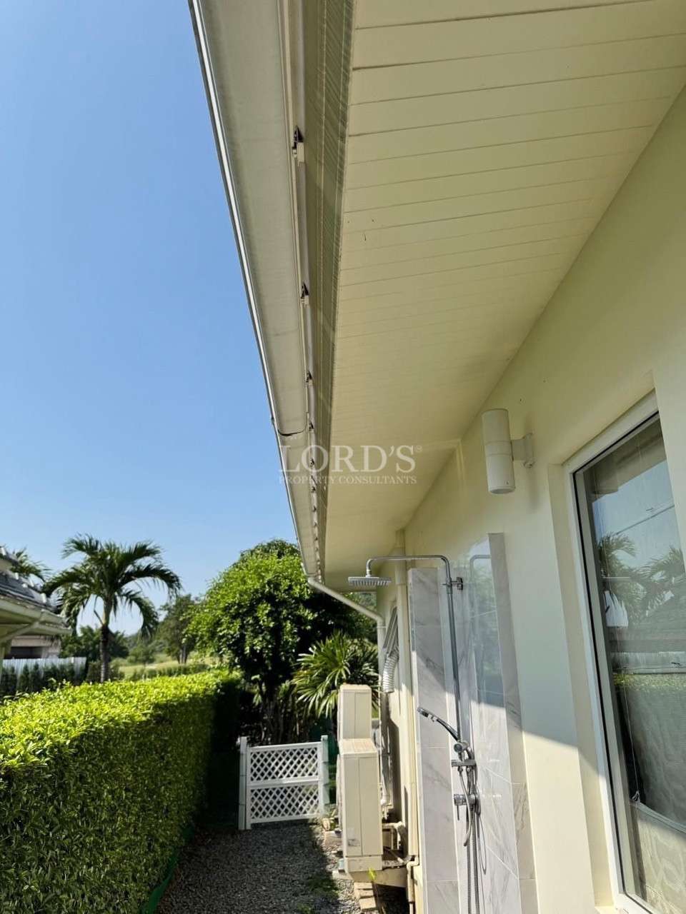 Side exterior of a house with a covered eave, outdoor shower fixture, window, trimmed hedge, and palm trees under a clear blue sky.