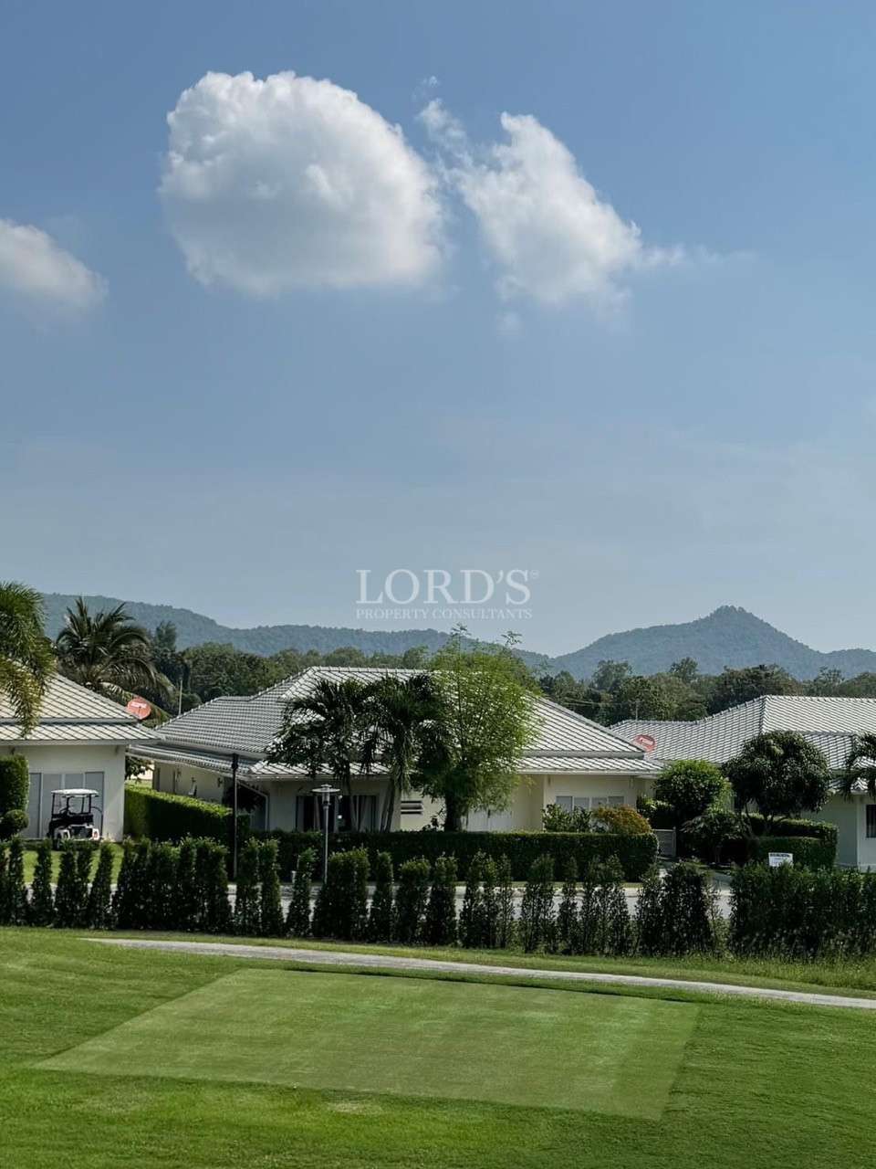 Single-story houses with light tiled roofs behind a hedge, set against green hills and a blue sky with scattered clouds.