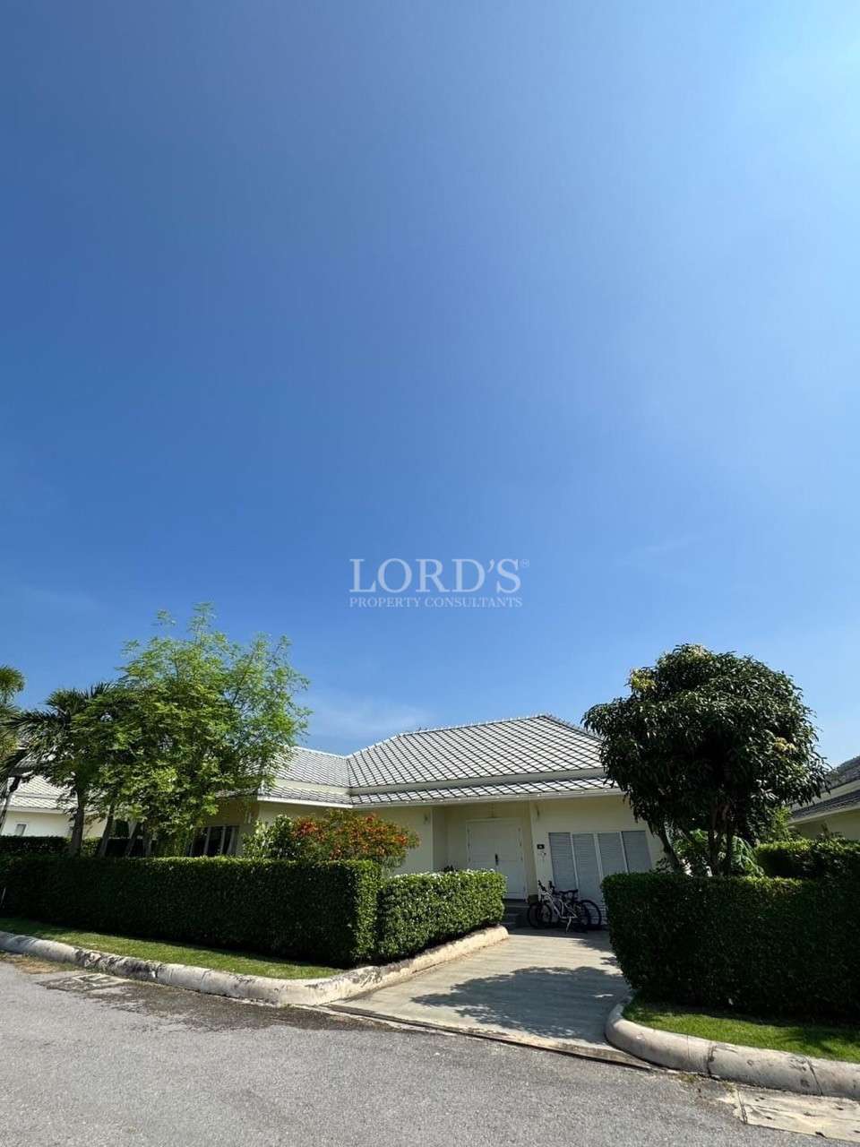 Single-story white house with a tiled roof, trimmed hedges, trees, and bicycles parked near the entrance under a clear blue sky.