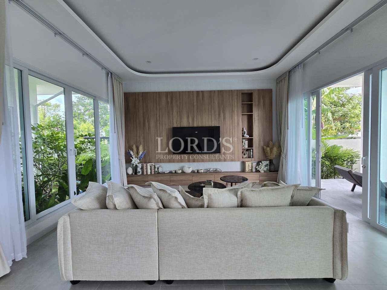 Modern living room with beige sofa, wood-paneled TV wall, and large windows overlooking greenery.