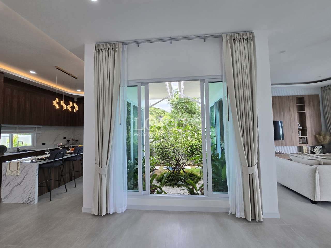 Large sliding glass window with curtains overlooking a lush indoor garden courtyard between kitchen and living areas.