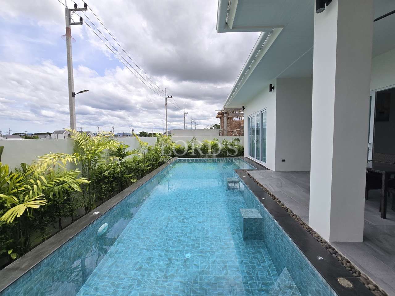 Narrow rectangular swimming pool beside a modern white house with sliding glass doors and tropical plants.