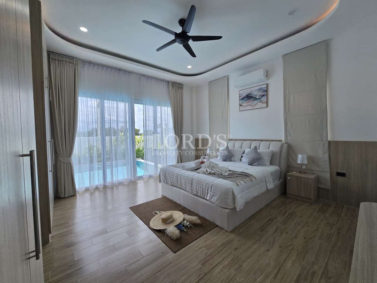 Modern bedroom with upholstered bed, wood flooring, ceiling fan, and sliding glass doors overlooking a pool.