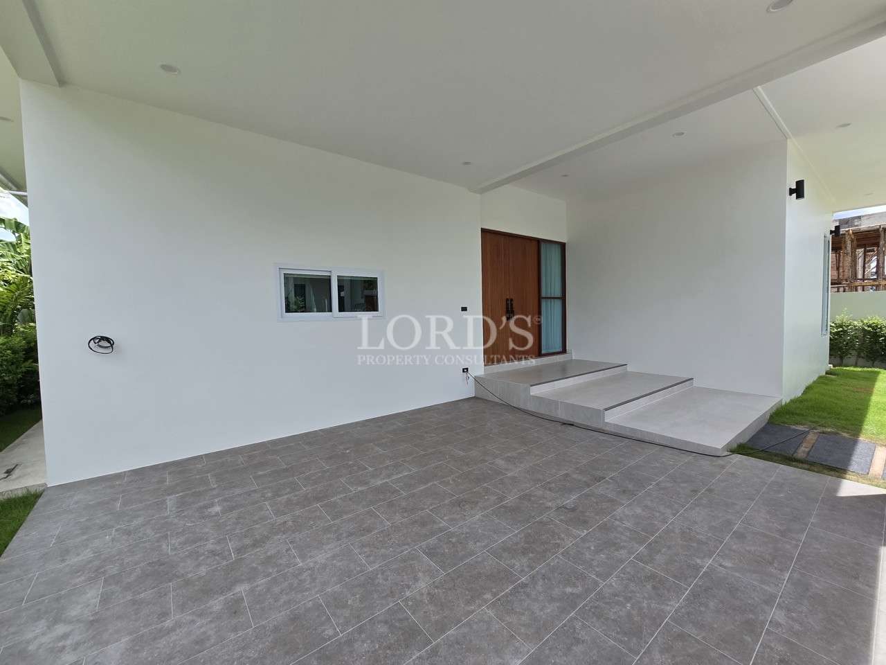 Covered outdoor patio area with tiled flooring, white walls, and steps leading to a wooden front door.