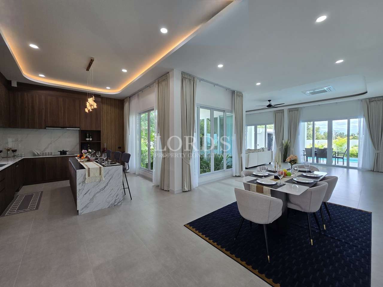 Bright open-plan kitchen and dining area with marble island, modern lighting, and large glass doors leading to a patio and pool.