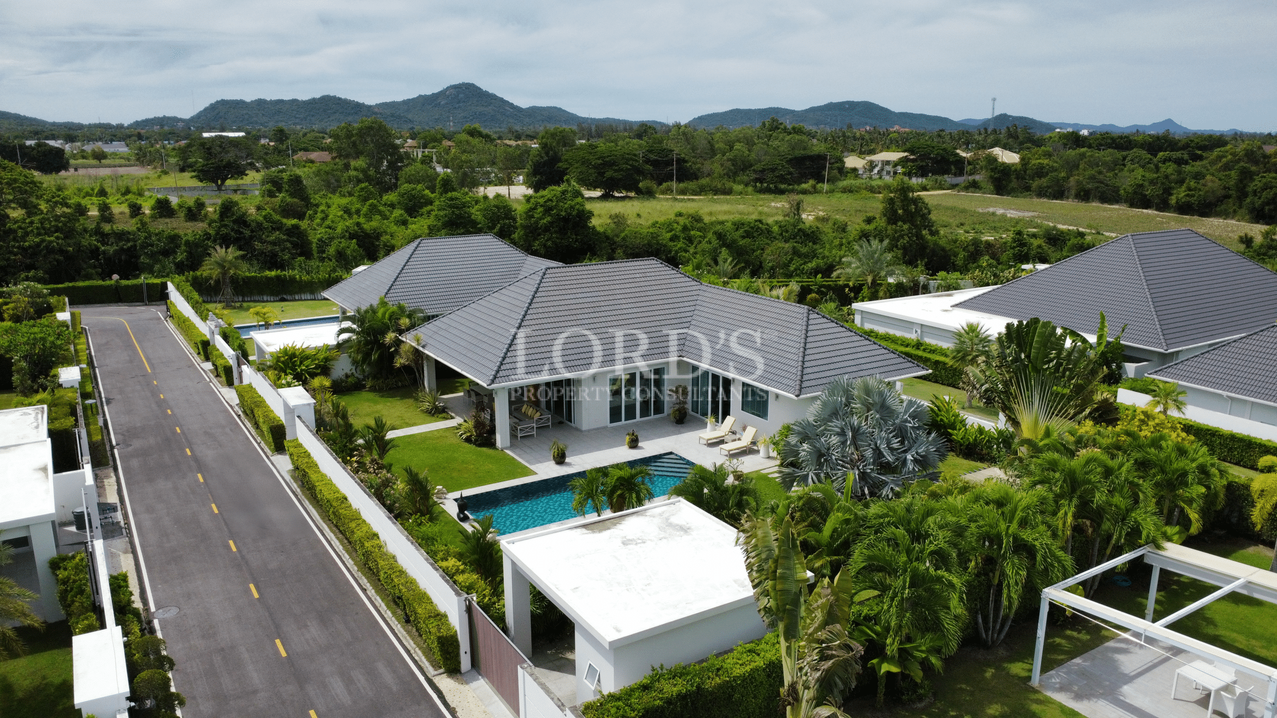 Aerial view of a modern pool villa with a gray tiled roof, private swimming pool, and landscaped garden near green hills.