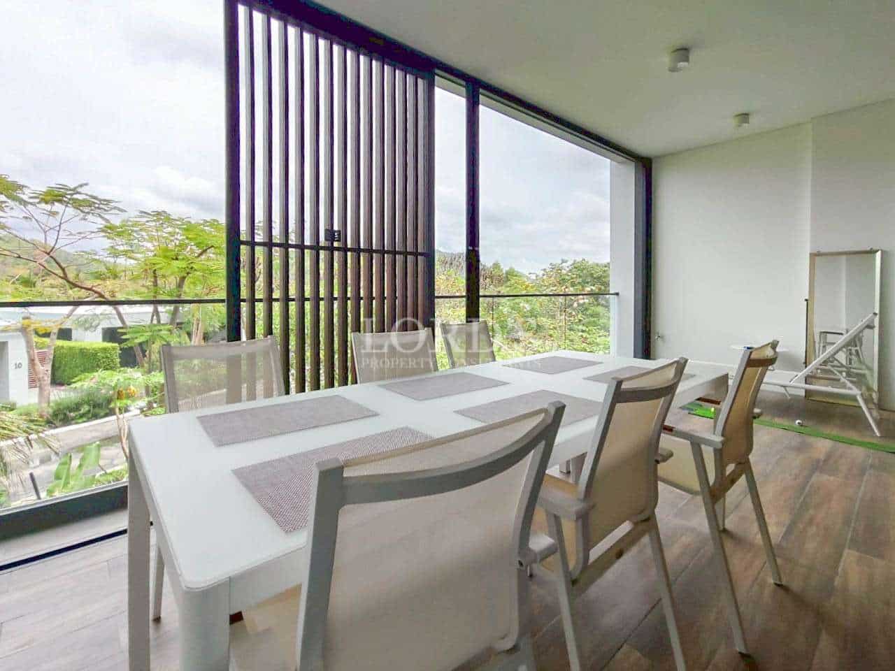 Outdoor balcony dining area with white table, chairs, and green garden view.