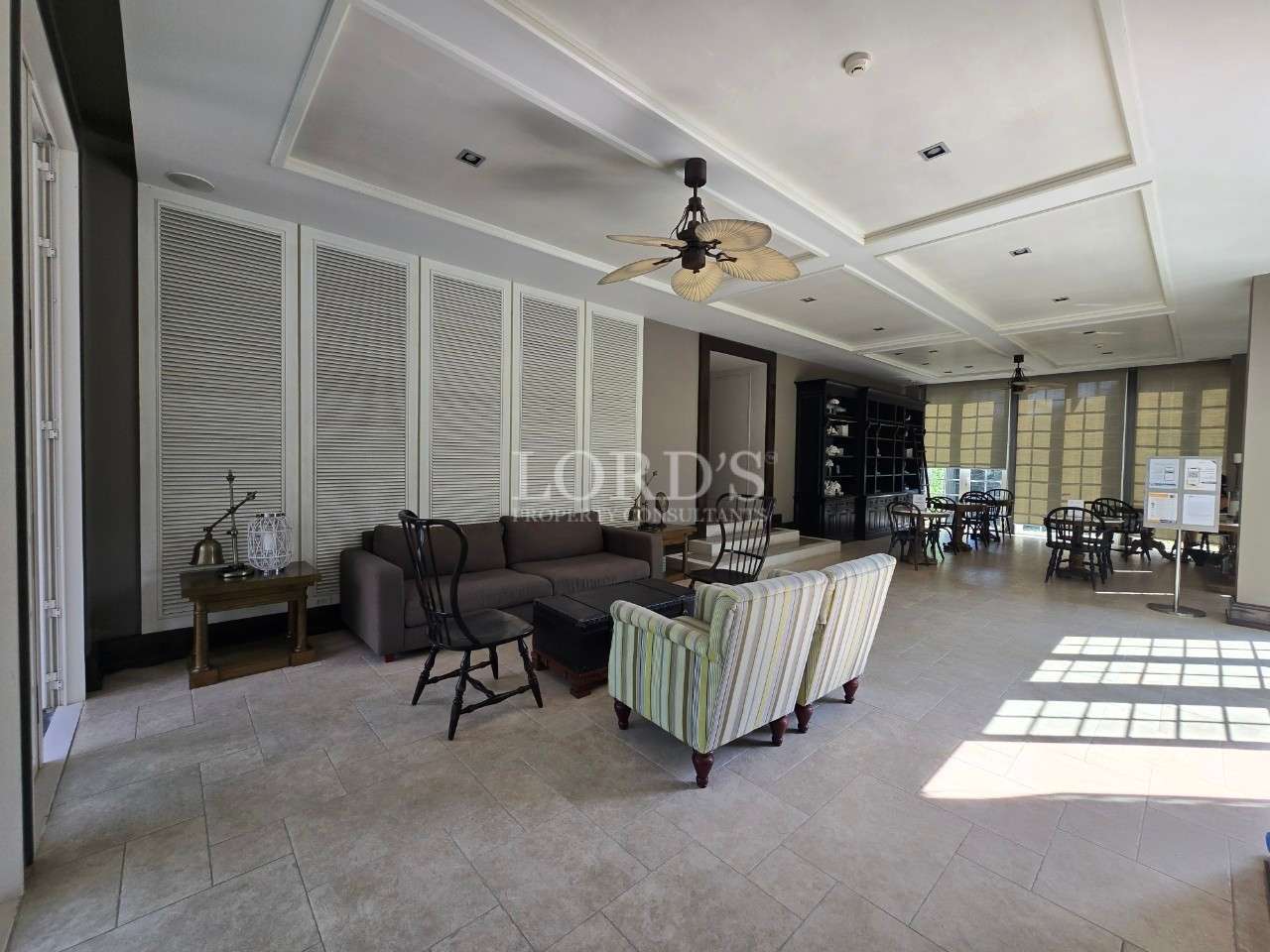 Spacious lounge area with sofas, armchairs, ceiling fans, and dining tables in a bright indoor common space.