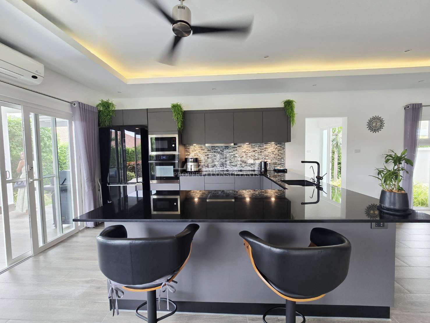 Modern open-plan kitchen with black island, bar stools, ceiling fan, and built-in appliances.