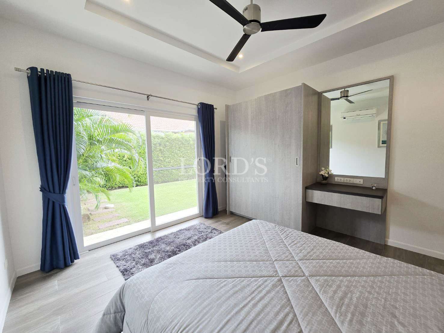 Bright bedroom with large glass doors, garden view, ceiling fan, wardrobe, and modern vanity.