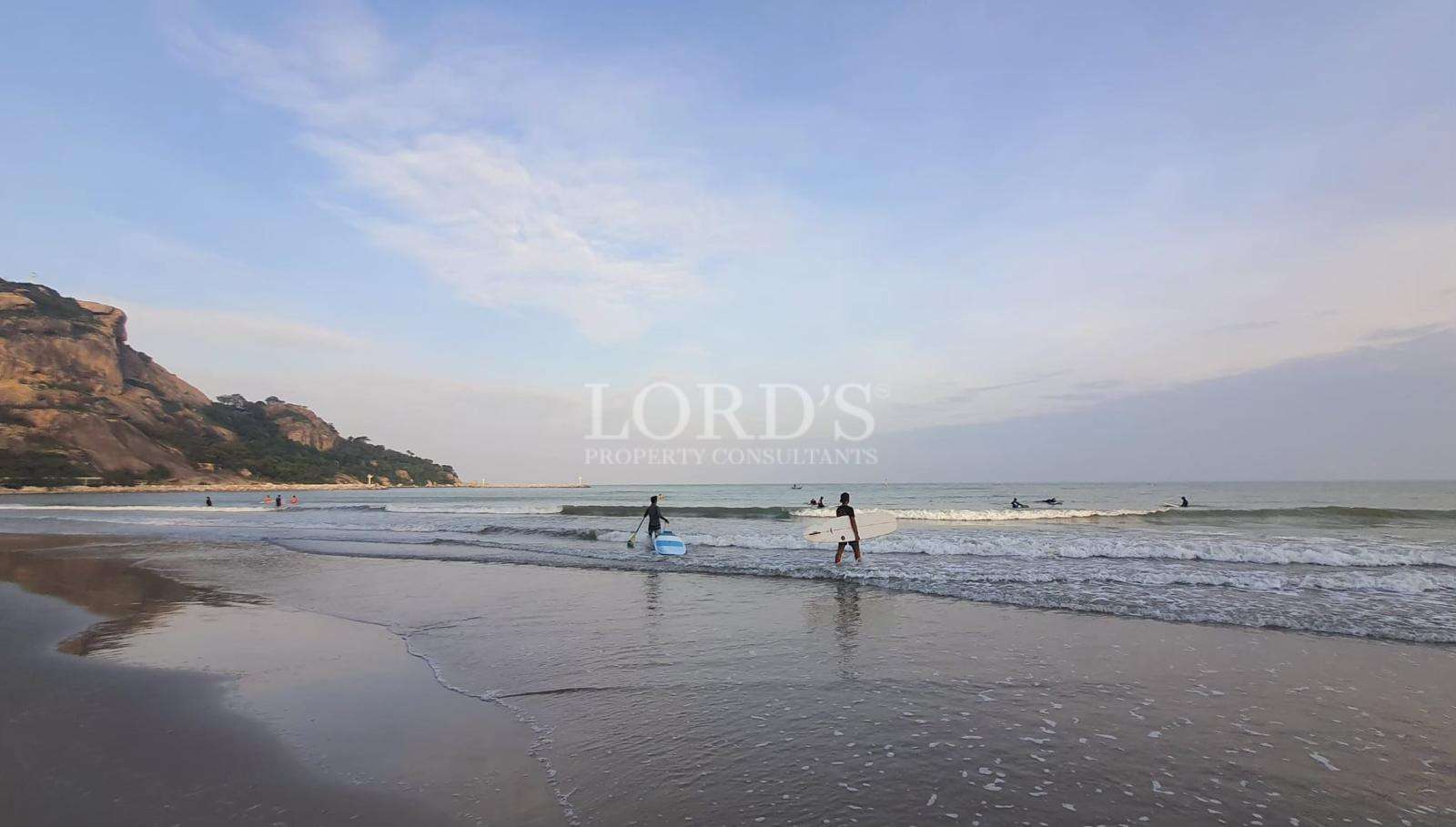 Surfers walking into the ocean at a sandy beach with gentle waves and rocky hills in the background.