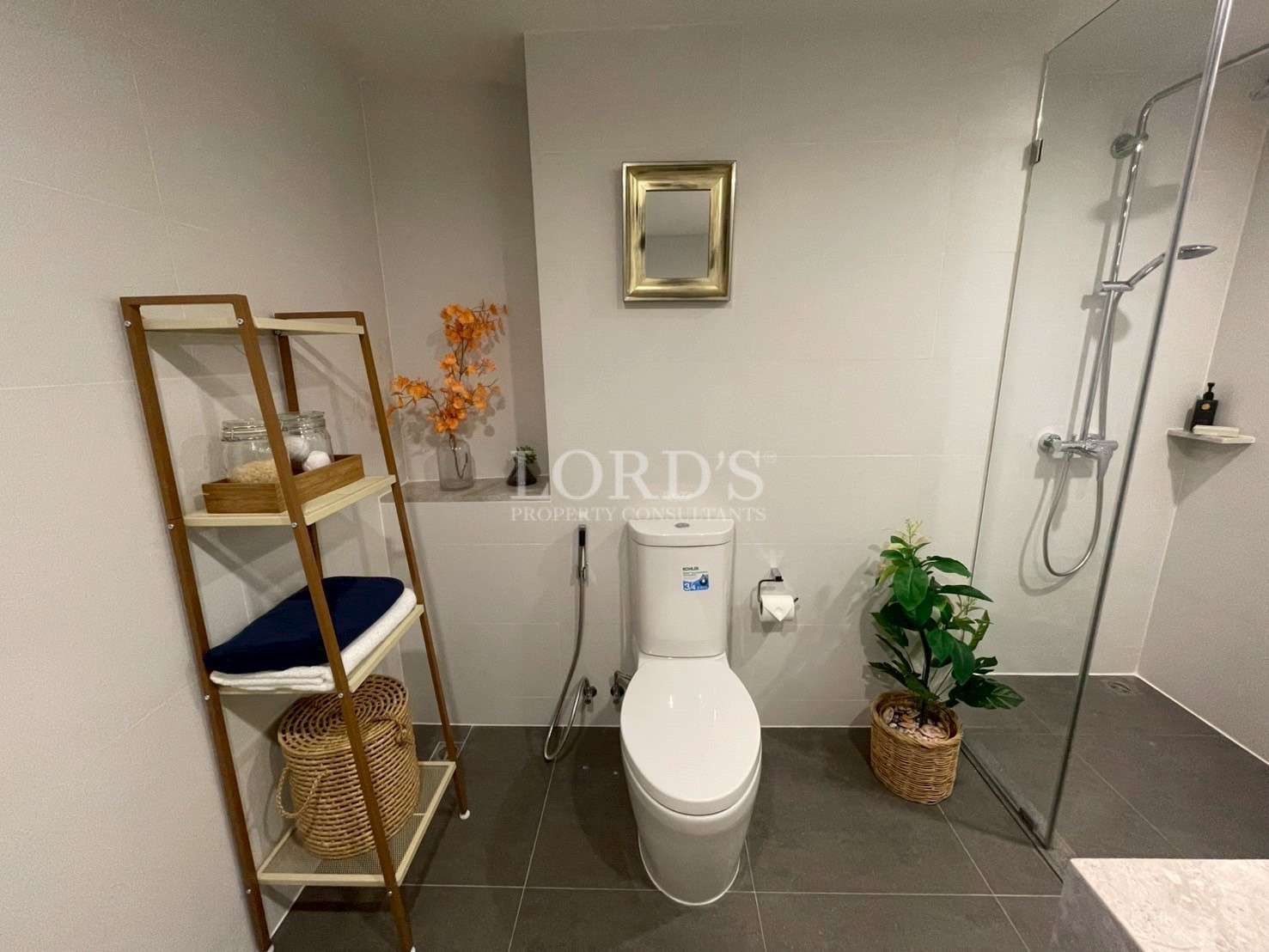 Modern bathroom with toilet, glass shower enclosure, wooden storage rack, and indoor plants.