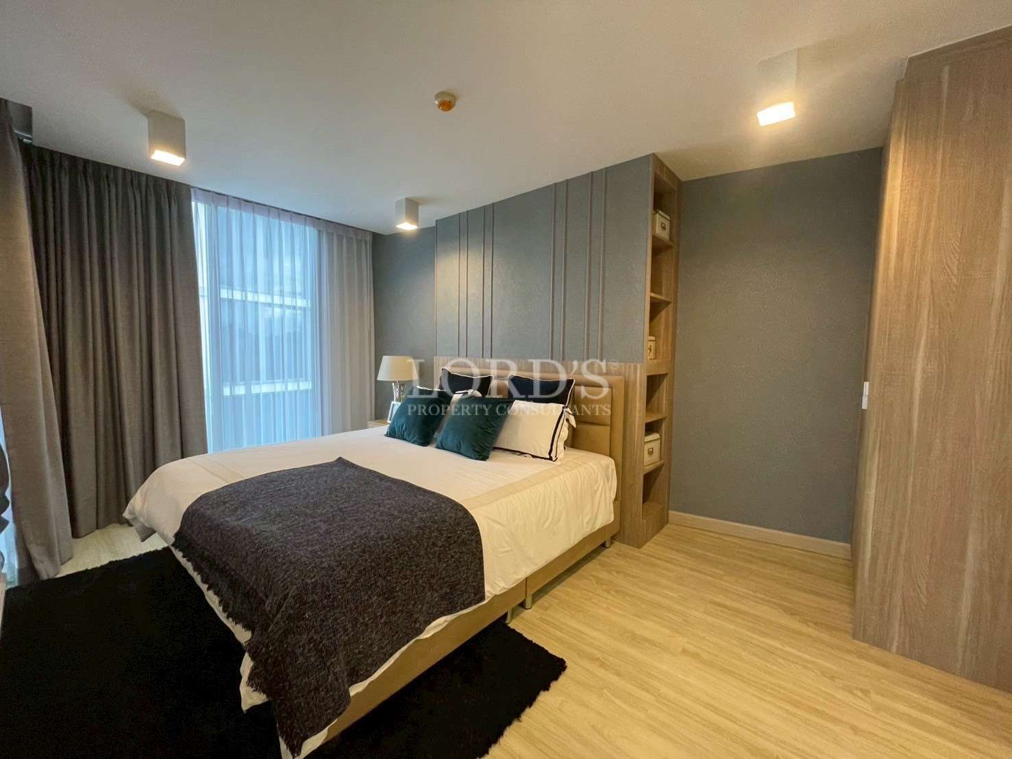 Modern bedroom with double bed, built-in shelving, wooden flooring, and large window curtains.