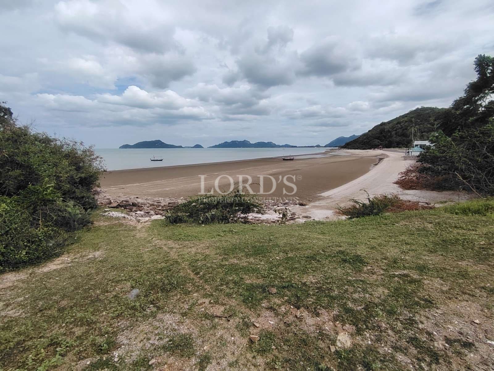 Wide sandy beach with calm sea, small boats offshore, and distant islands under a cloudy sky.
