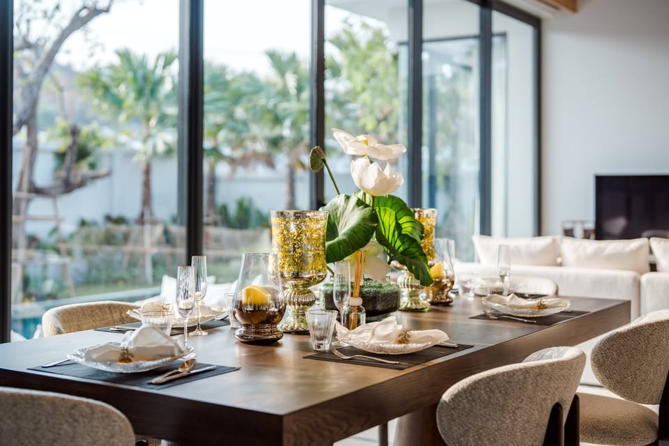 Modern dining table set with elegant tableware, floral centerpiece, and large glass windows overlooking a garden.