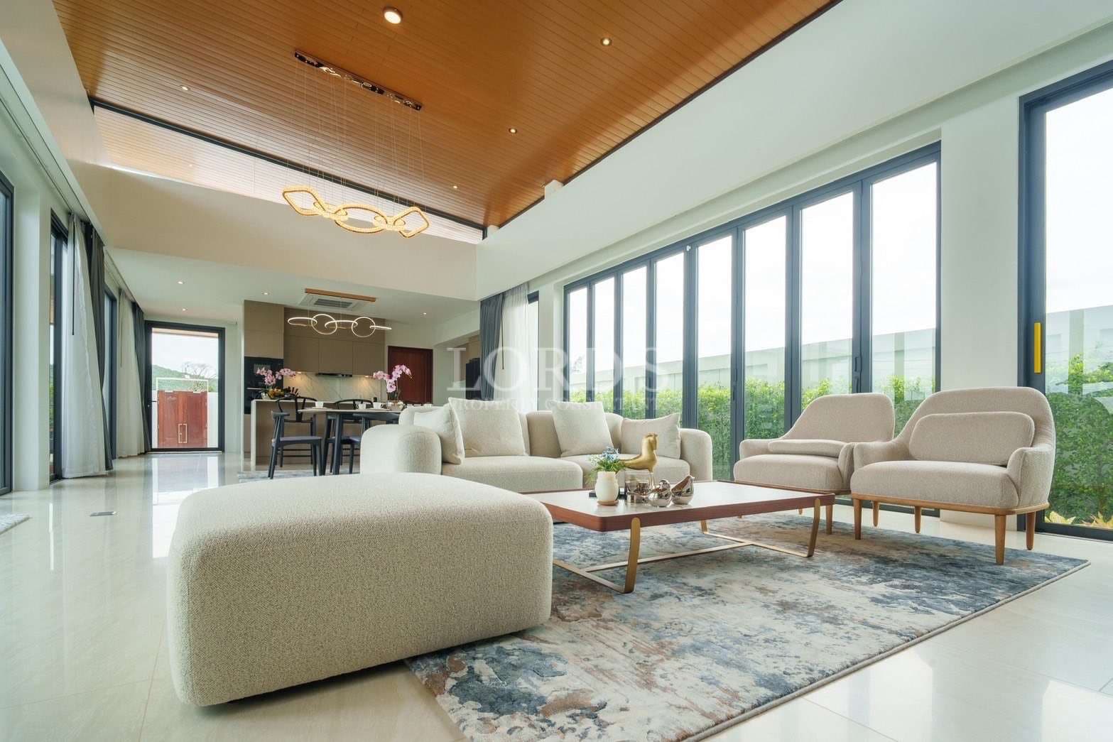 Luxury living room with high ceiling, wooden ceiling panels, modern furniture, and floor-to-ceiling windows.