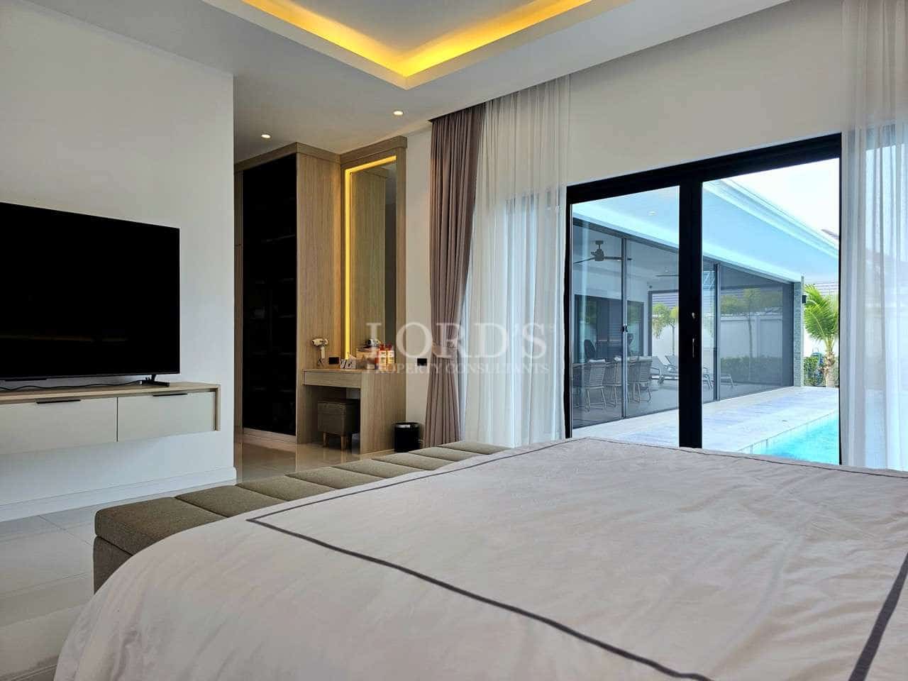 Modern bedroom interior with large bed, TV unit, vanity area, and sliding glass doors opening to a private pool