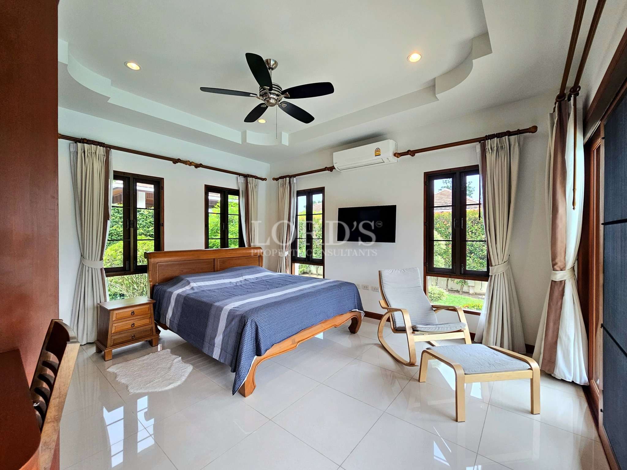 Bright villa bedroom with wooden bed, ceiling fan, large windows, and lounge chair