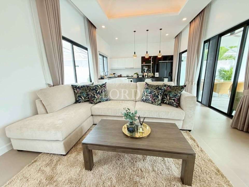 Modern living room with beige sectional sofa, wooden coffee table, large windows, and open-plan kitchen in the background.