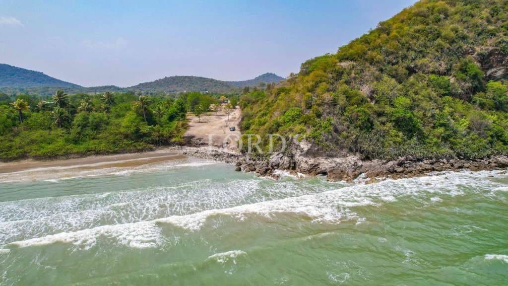 Aerial view of a secluded beach with gentle waves, rocky headland, and lush green hills in the background.