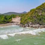 Luftaufnahme eines abgelegenen Strandes mit sanften Wellen, einer felsigen Landzunge und üppig grünen Hügeln im Hintergrund.