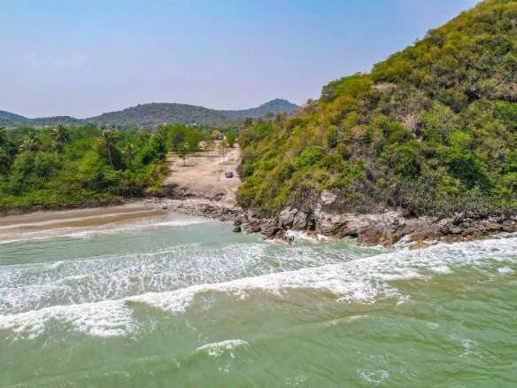 Aerial view of a secluded beach with gentle waves, rocky headland, and lush green hills in the background.