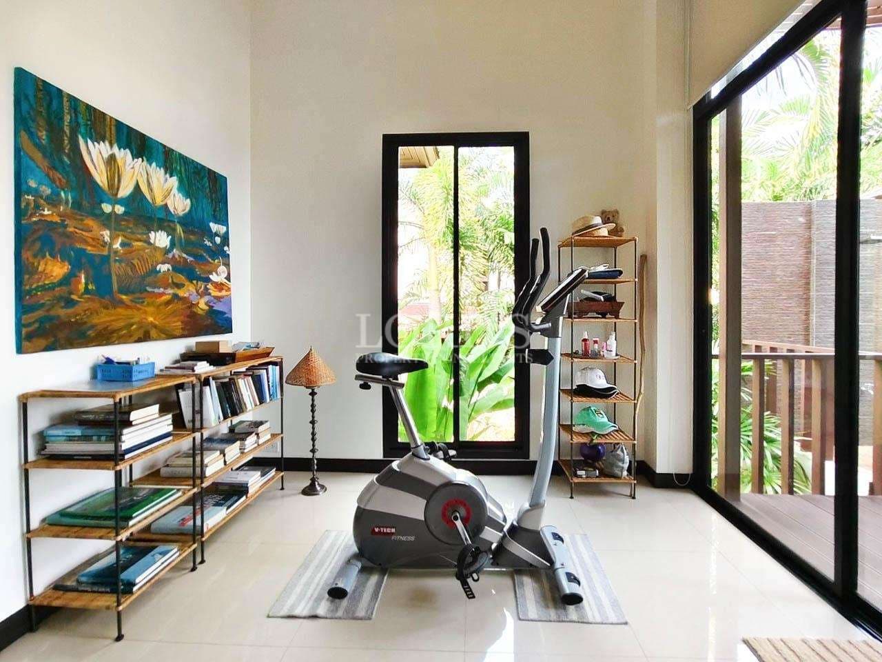Bright home gym room with exercise bike, bookshelf, artwork, and sliding glass doors overlooking tropical greenery.