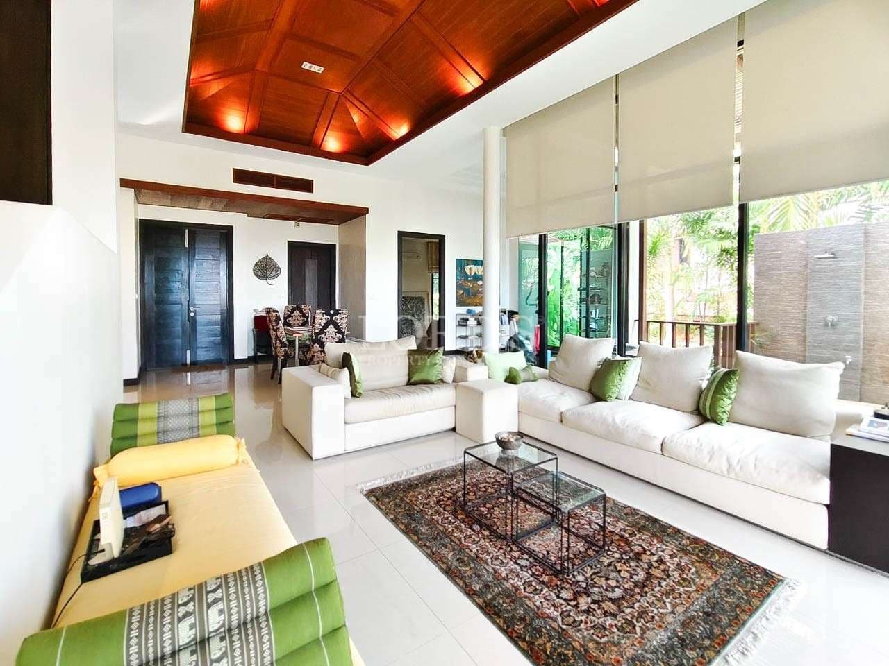Spacious luxury living room with white sofas, green accent cushions, wooden ceiling, and large glass windows opening to a tropical garden.