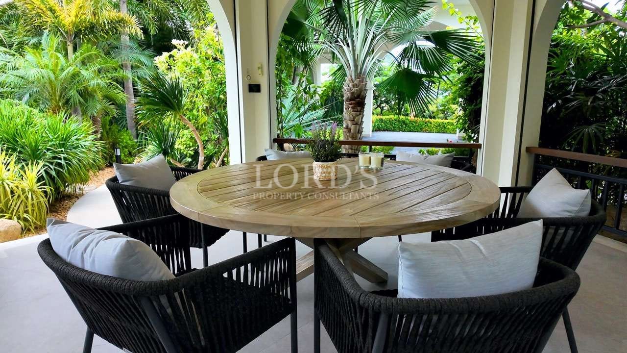 Outdoor patio dining area with round wooden table, wicker chairs, and lush tropical garden view.