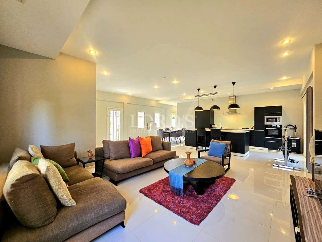 Modern open-plan living room with cozy sofa seating, central coffee table, recessed ceiling lights, and a contemporary black kitchen in the background.