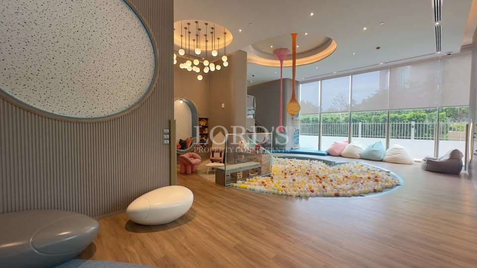 Modern indoor kids playroom with soft play area, ball pit, pastel seating, decorative ceiling lights, and large floor-to-ceiling windows in a luxury residential complex.